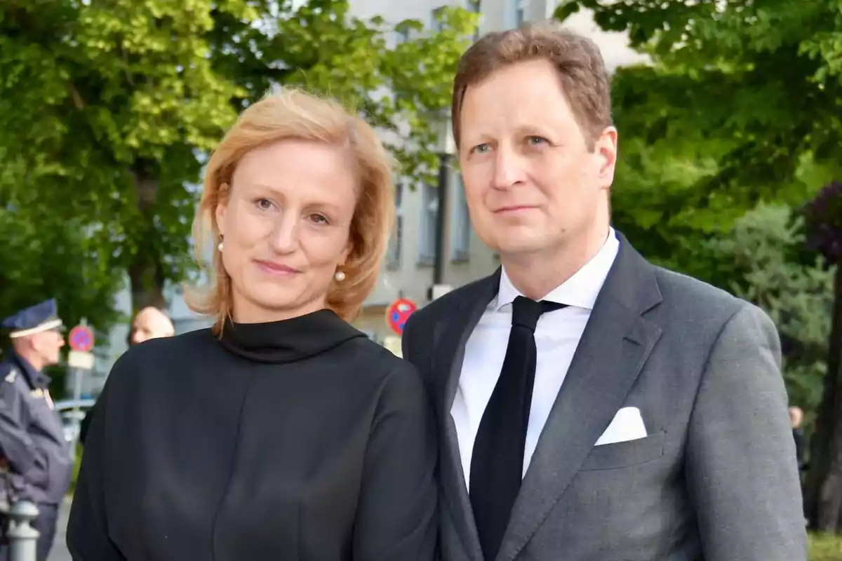 Una pareja vestida formalmente posa al aire libre con árboles y un edificio de fondo. Una pareja vestida formalmente posa al aire libre con árboles y un edificio de fondo.