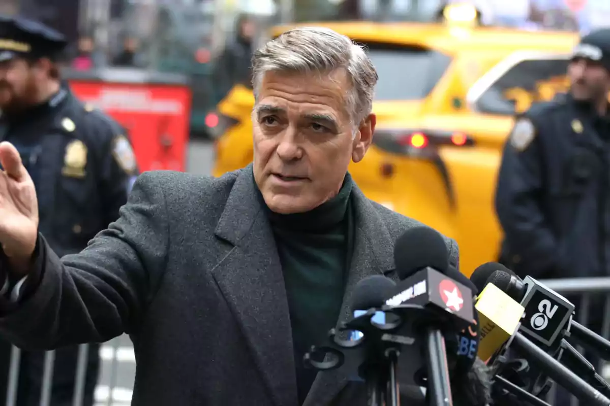 George Clooney con abrigo oscuro habla frente a varios micrófonos con un taxi amarillo y policías de fondo. George Clooney con abrigo oscuro habla frente a varios micrófonos con un taxi amarillo y policías de fondo.