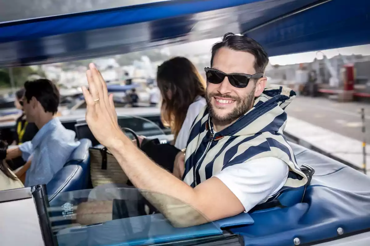 Hombre sonriente con gafas de sol y chaleco a rayas saludando desde un vehículo descapotable.