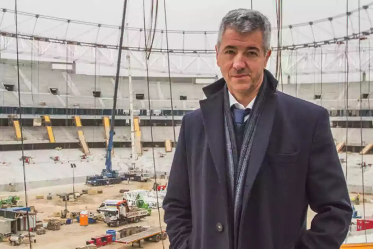 Hombre de pie con abrigo oscuro en un estadio en construcción.