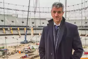 Hombre de pie con abrigo oscuro en un estadio en construcción.