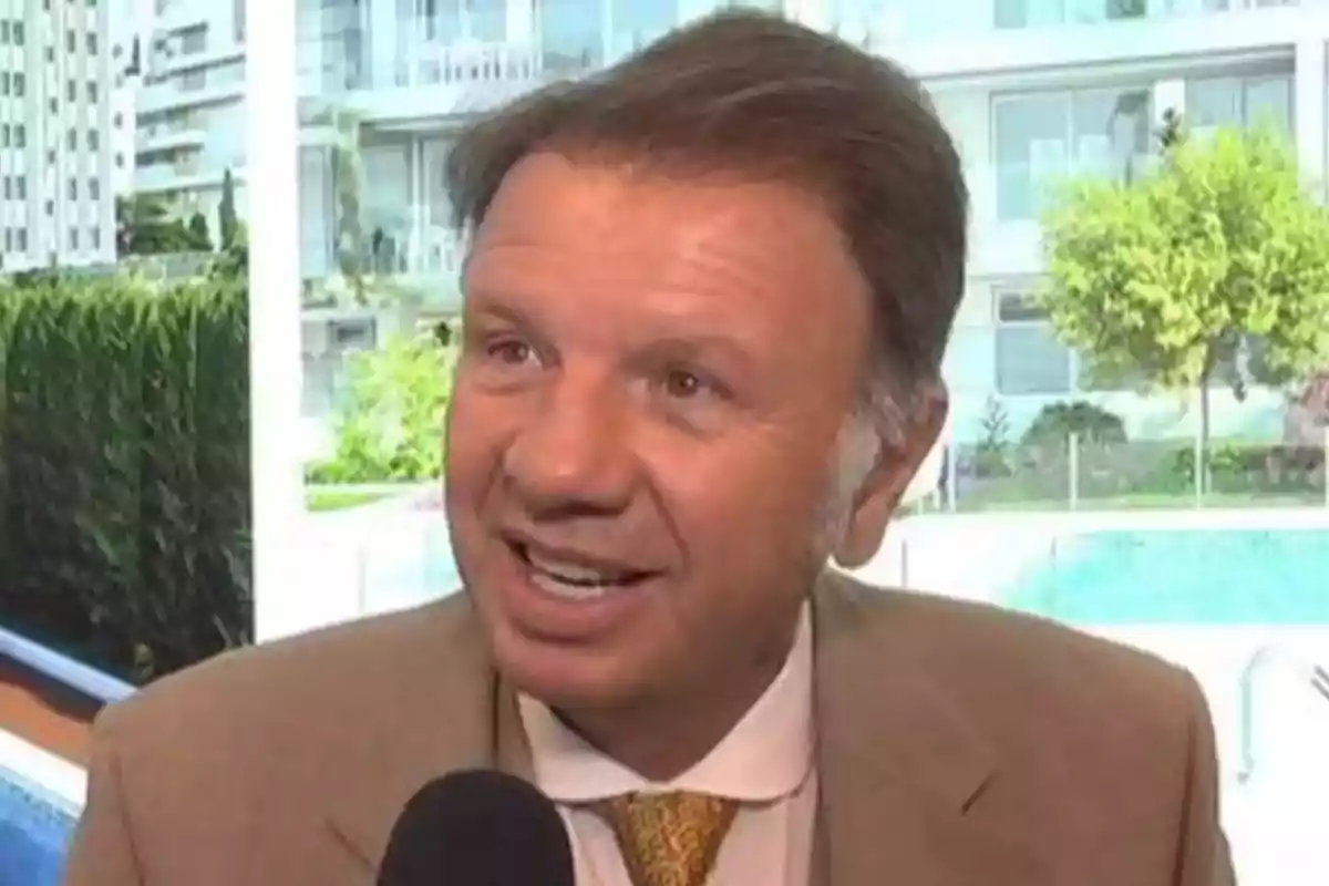 Hombre de traje claro sonriendo mientras sostiene un micrófono con fondo de edificios y vegetación
