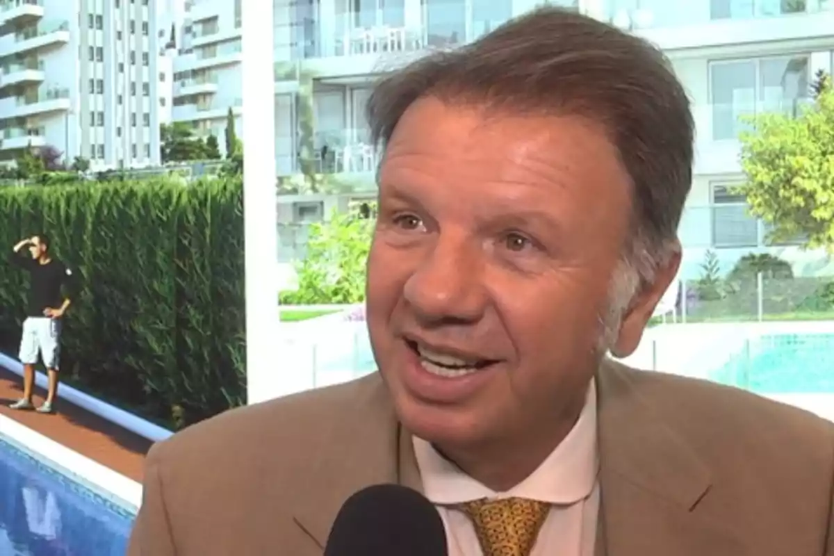 Un hombre sonriente con traje y corbata es entrevistado frente a un fondo de edificios y una piscina.