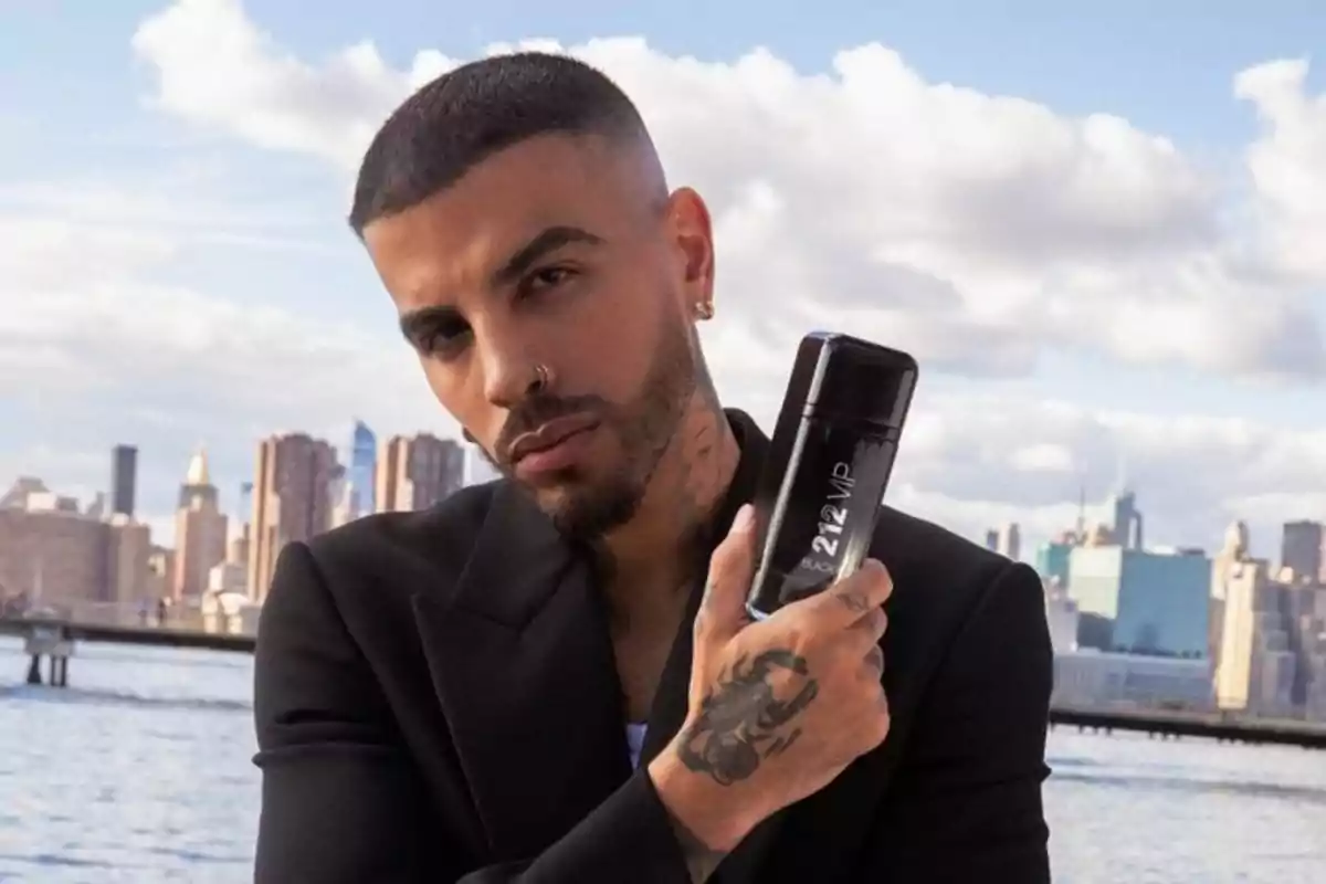 Hombre con barba y tatuajes sosteniendo una botella de perfume 212 VIP Black frente a un paisaje urbano con edificios y agua.