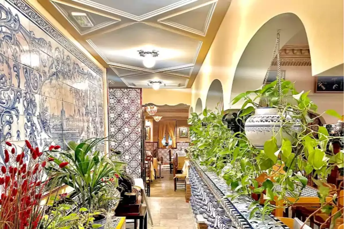 Un pasillo decorado con azulejos azules y blancos, plantas verdes colgantes y macetas, con mesas y sillas al fondo.