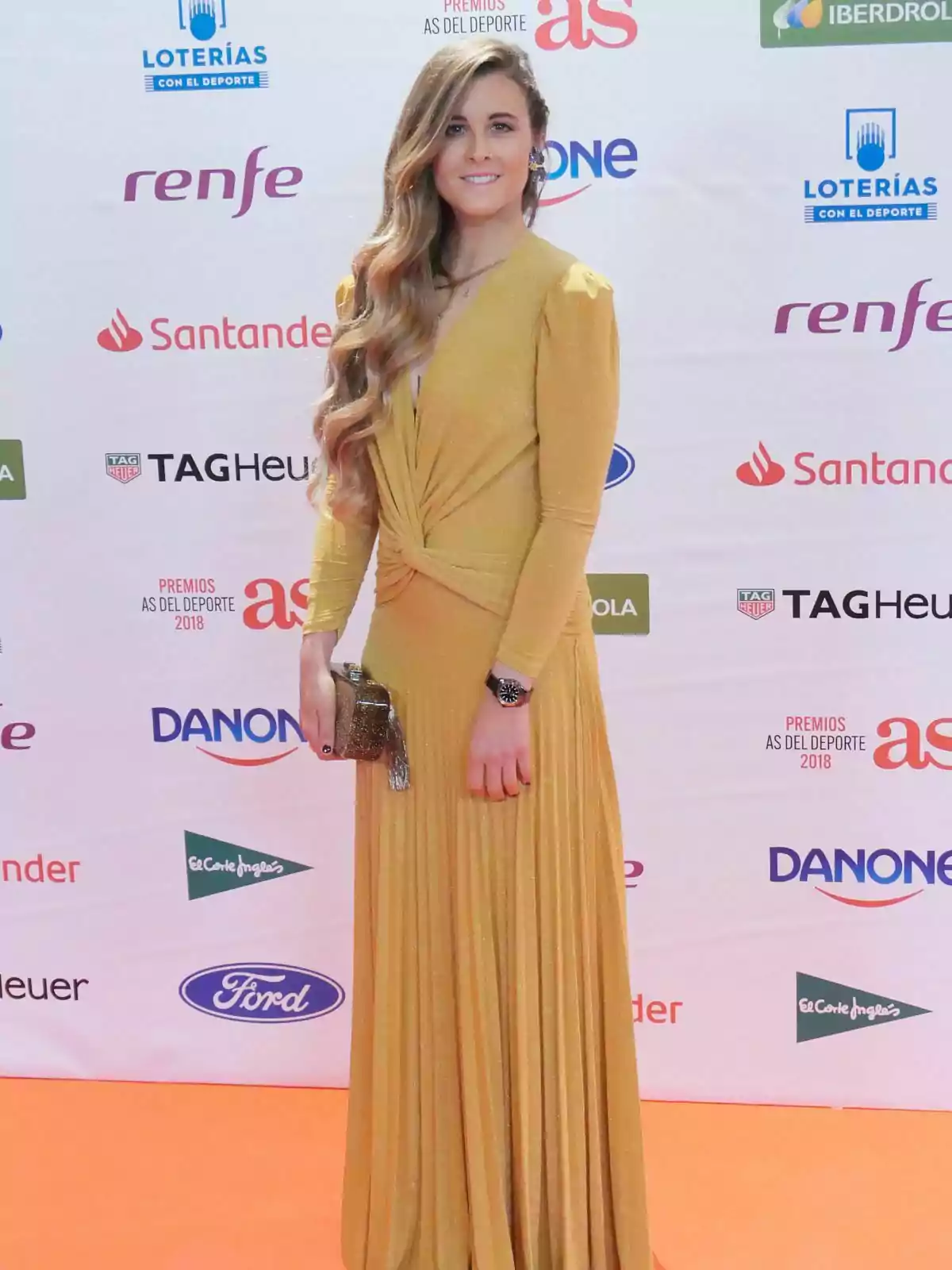 Gisela Pulido con un vestido largo dorado posa en una alfombra naranja frente a un fondo con logotipos de varias marcas.