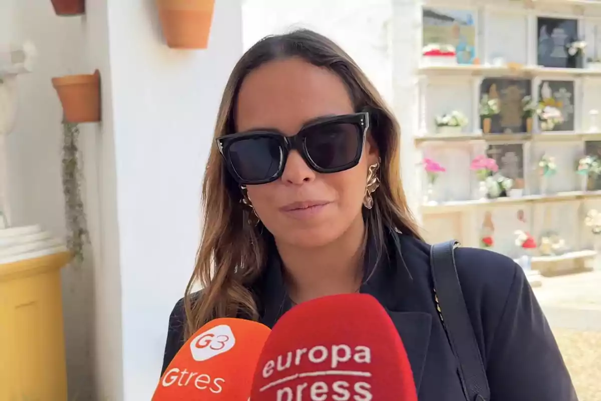 Gloria Camila con gafas de sol negras y chaqueta oscura responde preguntas frente a micrófonos de prensa en un cementerio Gloria Camila con gafas de sol negras y chaqueta oscura responde preguntas frente a micrófonos de prensa en un cementerio