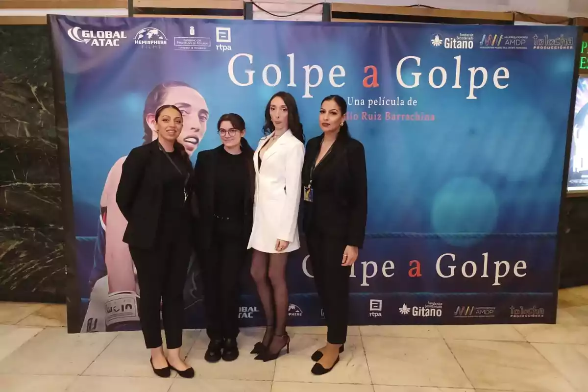 Un grupo de cuatro personas posa frente a un cartel promocional de la película "Golpe a Golpe".