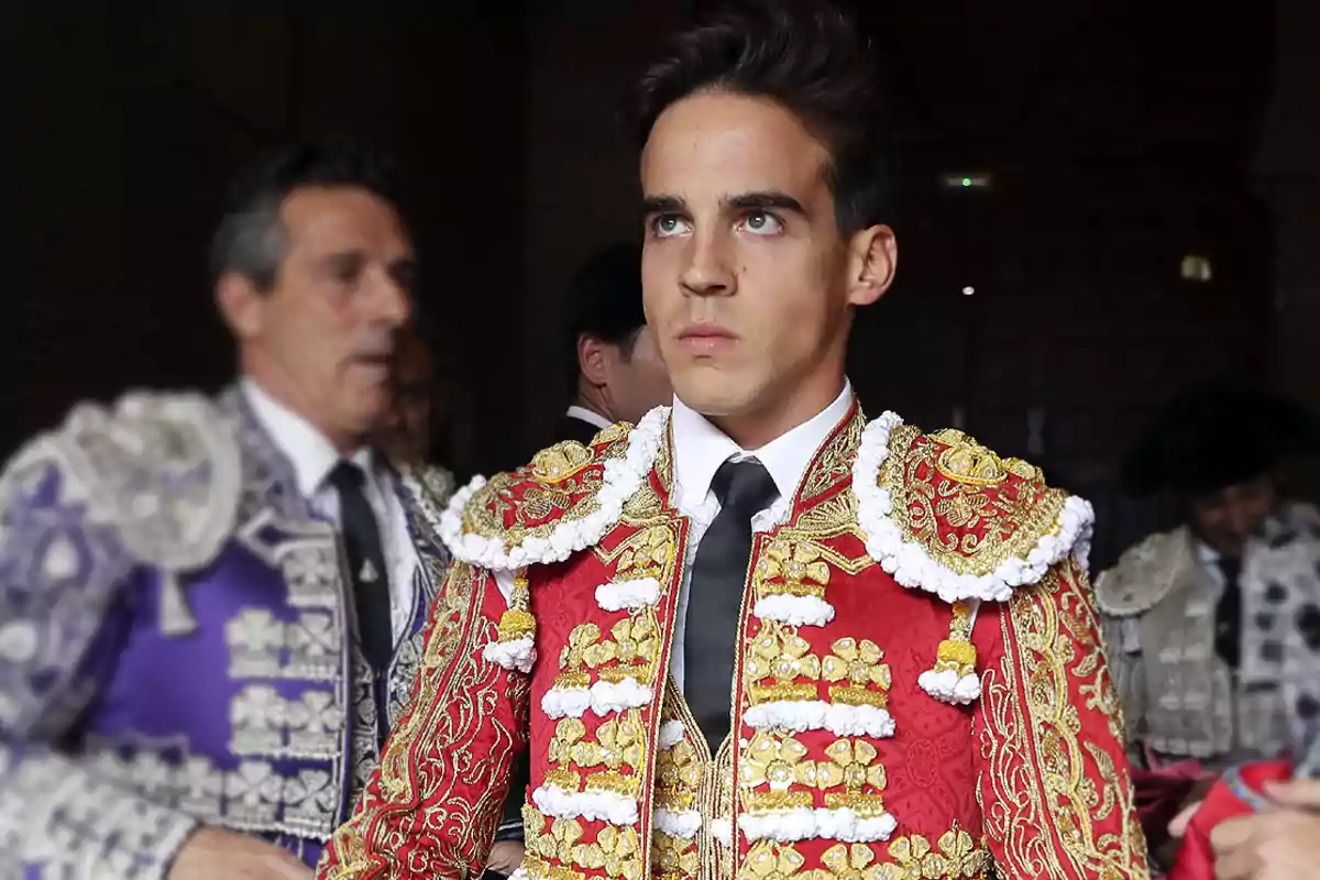 Un torero joven con traje de luces rojo y dorado, acompañado por otros toreros en el fondo.