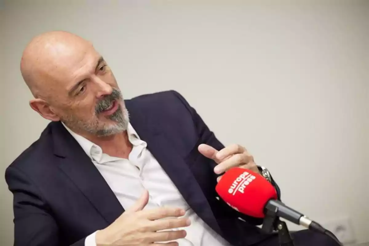Un hombre calvo con barba está hablando mientras sostiene un micrófono rojo de Europa Press. Un hombre calvo con barba está hablando mientras sostiene un micrófono rojo de Europa Press.