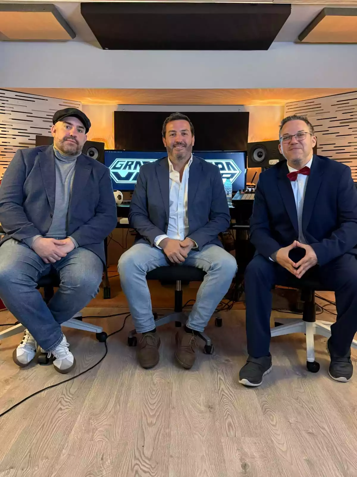 Tres hombres sentados en sillas de oficina en un estudio de grabación, todos vestidos con saco y uno de ellos lleva moño rojo Tres hombres sentados en sillas de oficina en un estudio de grabación, todos vestidos con saco y uno de ellos lleva moño rojo