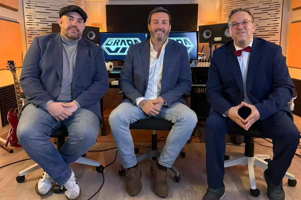 Tres hombres sentados en sillas de oficina dentro de un estudio de grabación con guitarras y equipo de audio al fondo