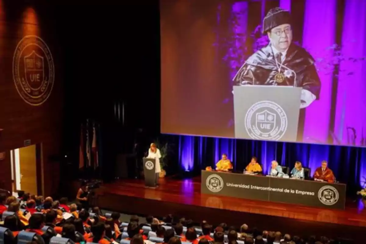 Auditorio lleno de personas asistiendo a una ceremonia académica en la Universidad Intercontinental de la Empresa con una mesa de autoridades en el escenario y una gran pantalla proyectando a un orador Auditorio lleno de personas asistiendo a una ceremonia académica en la Universidad Intercontinental de la Empresa con una mesa de autoridades en el escenario y una gran pantalla proyectando a un orador