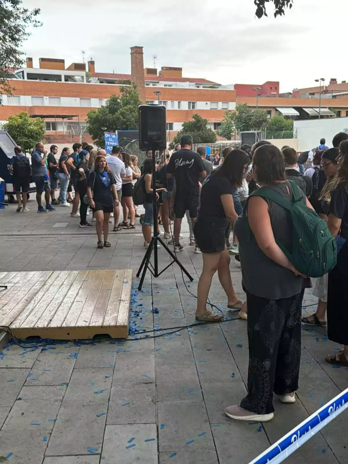 Un grupo de personas se encuentra reunido en una plaza al aire libre, algunas de ellas están haciendo fila cerca de una tarima de madera, mientras que otras conversan entre sí; en el suelo hay confeti azul esparcido y al fondo se observan edificios residenciales.