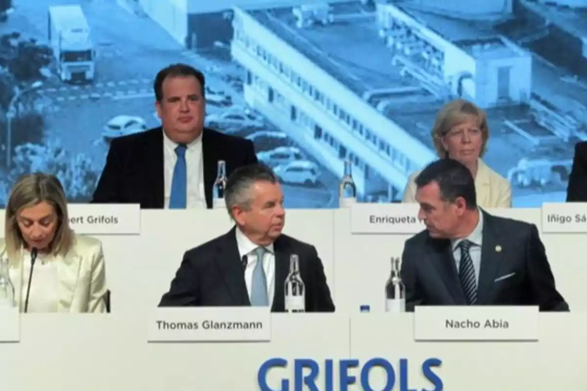 Un grupo de personas sentadas en una mesa de conferencia con letreros de nombres frente a ellos y una imagen de fondo de un edificio industrial.