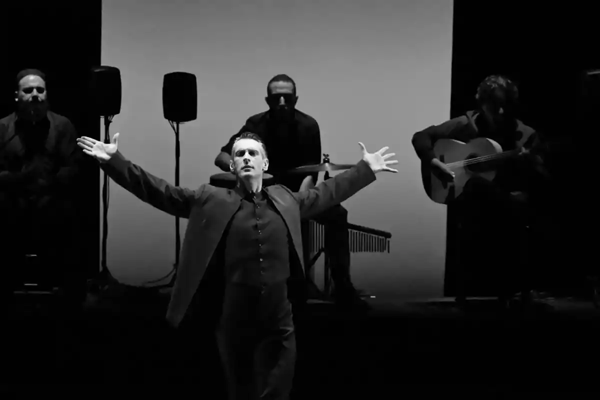 Un hombre bailando flamenco con los brazos extendidos, acompañado por tres músicos en un escenario oscuro.