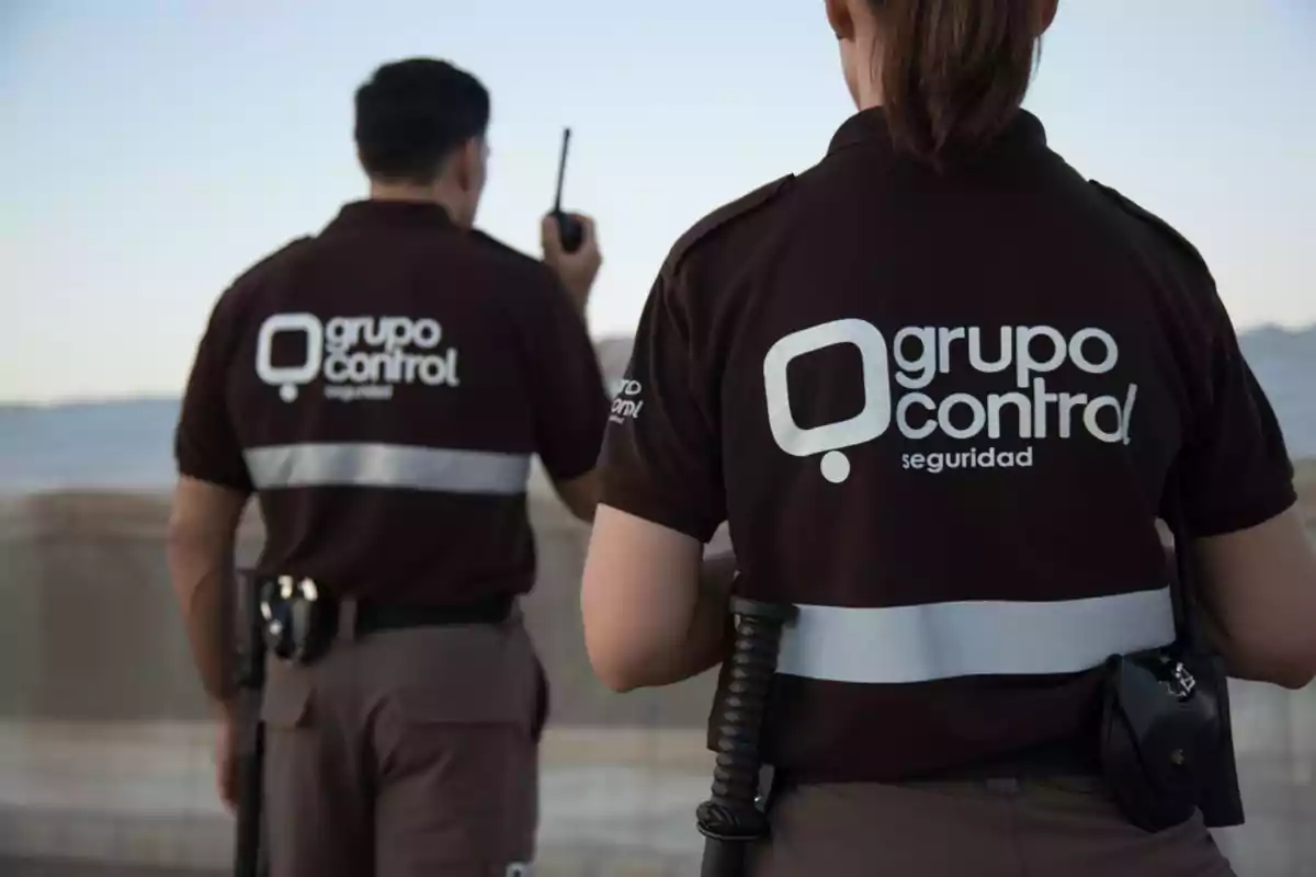 Dos guardias de seguridad de espaldas con uniformes marrones y logotipo de "Grupo Control" en la parte trasera, uno de ellos sostiene un walkie-talkie.