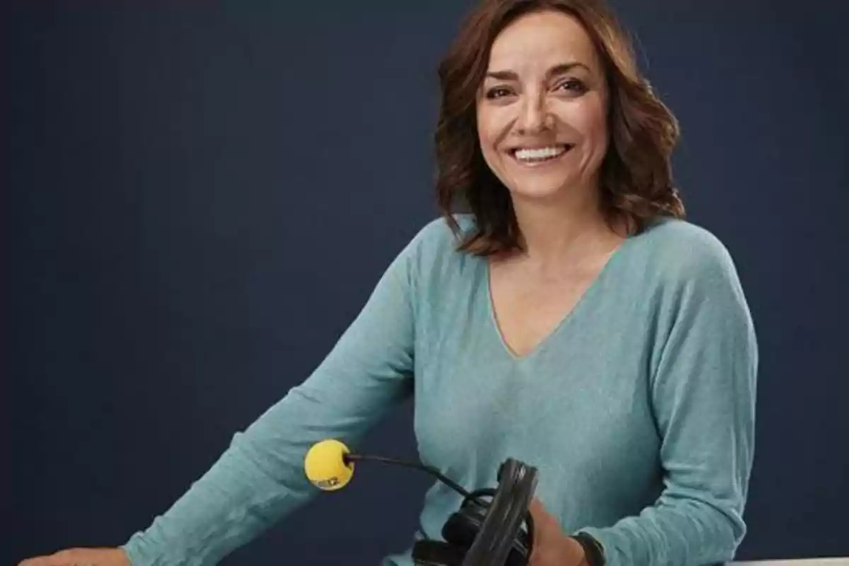 Mujer sonriente con cabello castaño y suelto, vestida con blusa azul claro, sostiene unos auriculares con micrófono de esponja amarilla frente a un fondo azul oscuro Mujer sonriente con cabello castaño y suelto, vestida con blusa azul claro, sostiene unos auriculares con micrófono de esponja amarilla frente a un fondo azul oscuro
