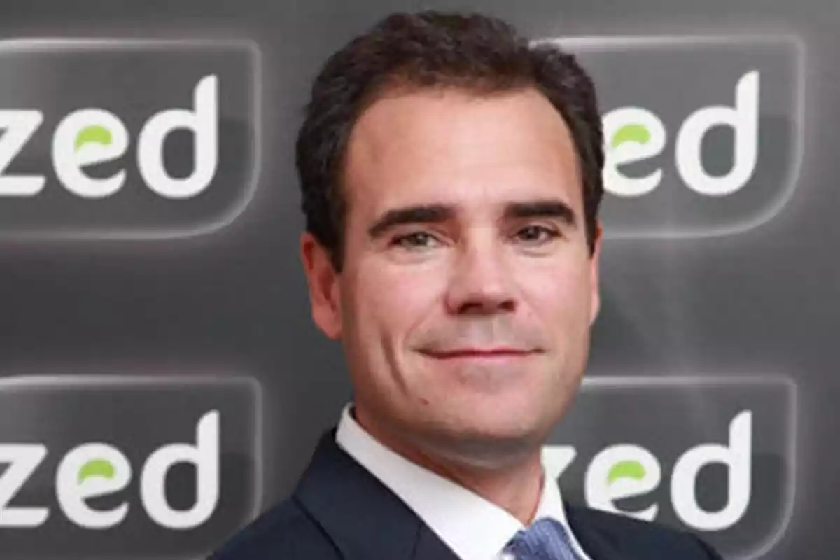 Hombre de cabello oscuro con traje y corbata posando frente a un fondo con el logo de la palabra zed repetido Hombre de cabello oscuro con traje y corbata posando frente a un fondo con el logo de la palabra zed repetido