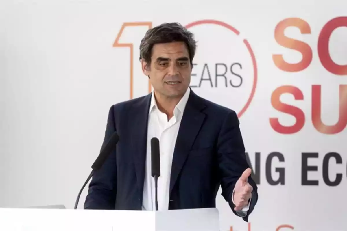 Un hombre en traje oscuro y camisa blanca habla en un podio con micrófonos frente a un fondo con texto parcialmente visible. Un hombre en traje oscuro y camisa blanca habla en un podio con micrófonos frente a un fondo con texto parcialmente visible.