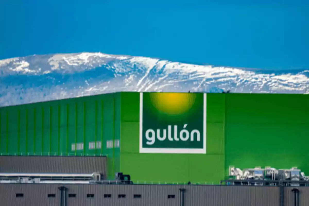 Edificio industrial verde con el logo de Gullón al frente de una montaña nevada bajo un cielo azul.