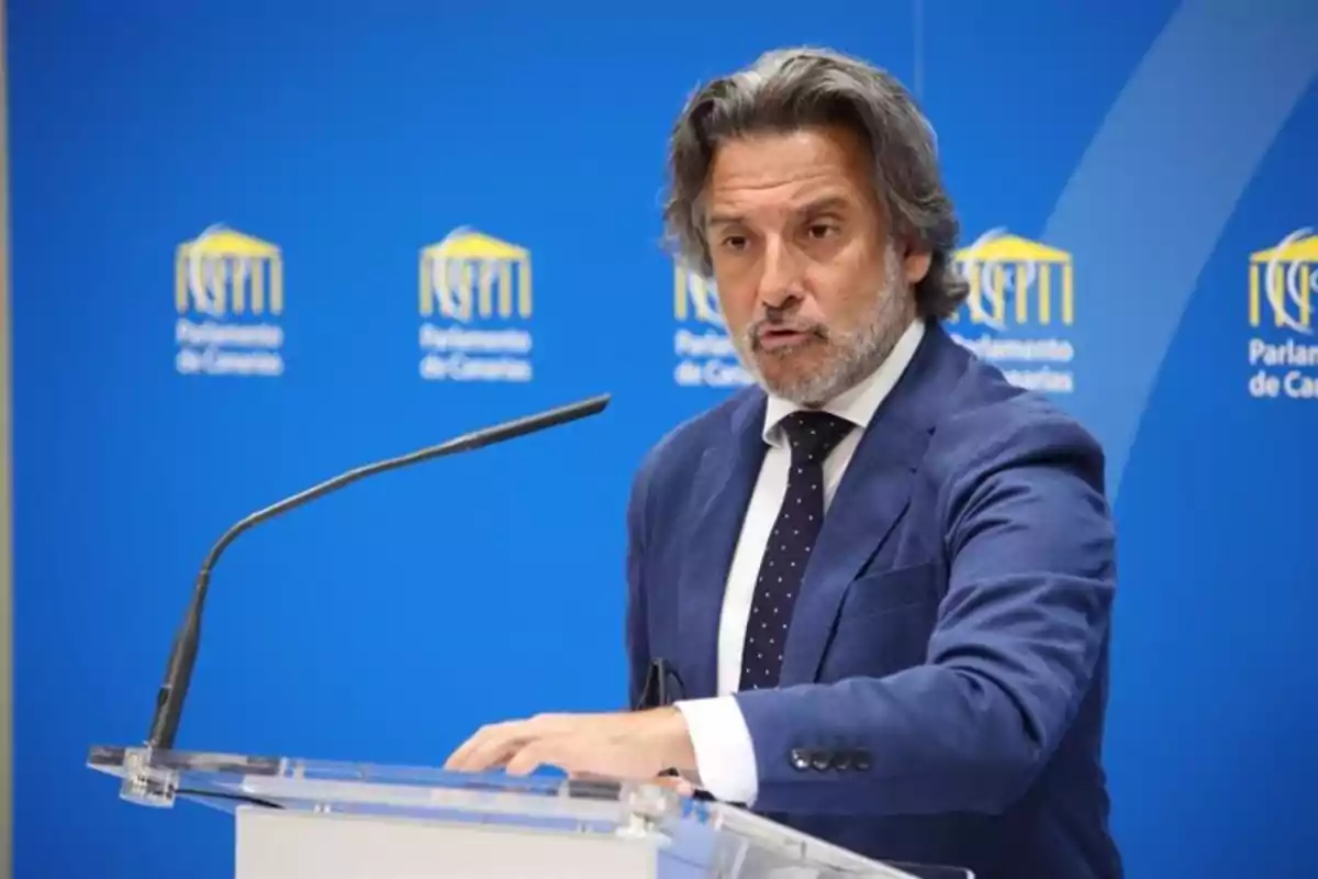 Un hombre de traje azul y corbata habla en un podio frente a un fondo azul con el logotipo del Parlamento de Canarias. Un hombre de traje azul y corbata habla en un podio frente a un fondo azul con el logotipo del Parlamento de Canarias.