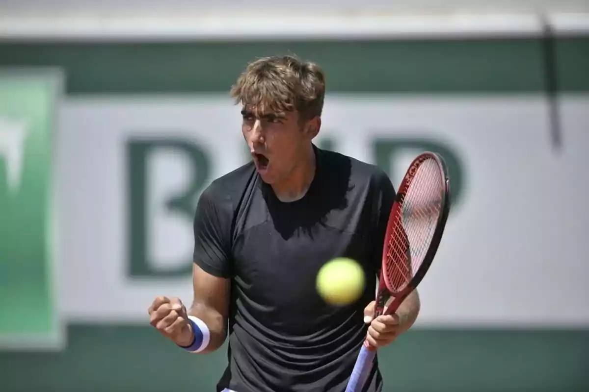 Joven tenista celebrando con gesto de emoción mientras sostiene una raqueta y una pelota de tenis flota frente a él