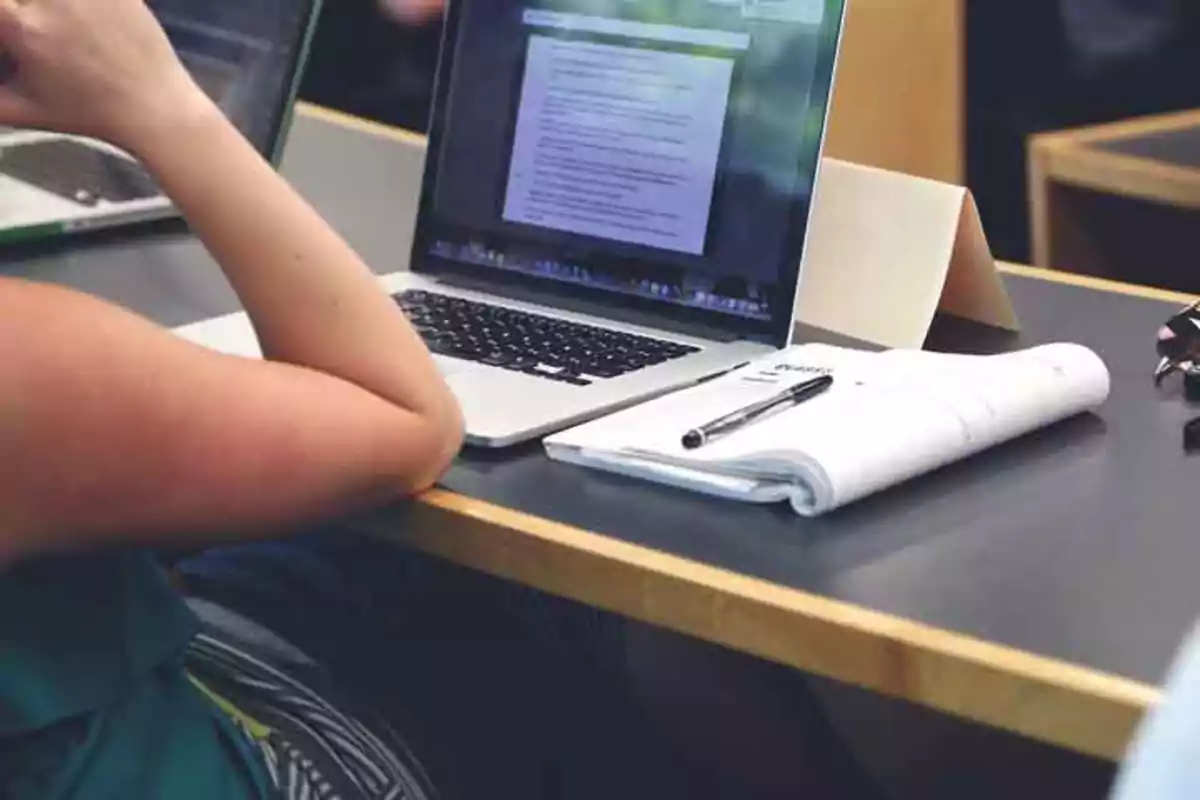 Persona trabajando en una computadora portátil con un cuaderno y un bolígrafo sobre la mesa.