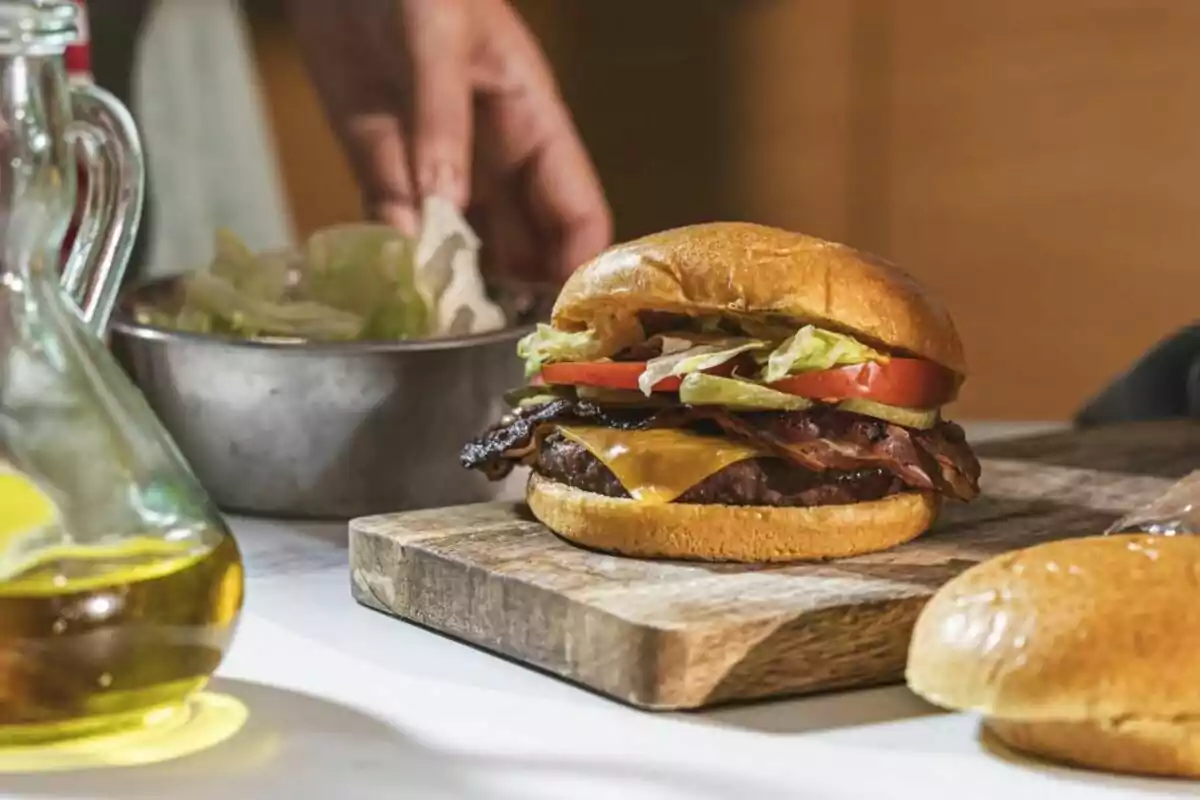 Hamburguesa con queso, tocino, lechuga y jitomate sobre una tabla de madera con una mano al fondo preparando ingredientes Hamburguesa con queso, tocino, lechuga y jitomate sobre una tabla de madera con una mano al fondo preparando ingredientes