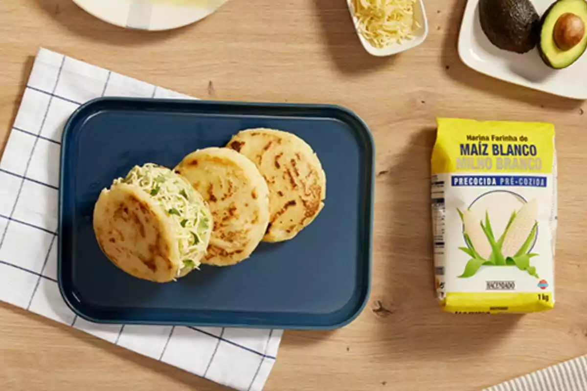 Tres arepas sobre un plato azul acompañadas de ingredientes como aguacate, queso rallado y harina de maíz blanco. Tres arepas sobre un plato azul acompañadas de ingredientes como aguacate, queso rallado y harina de maíz blanco.