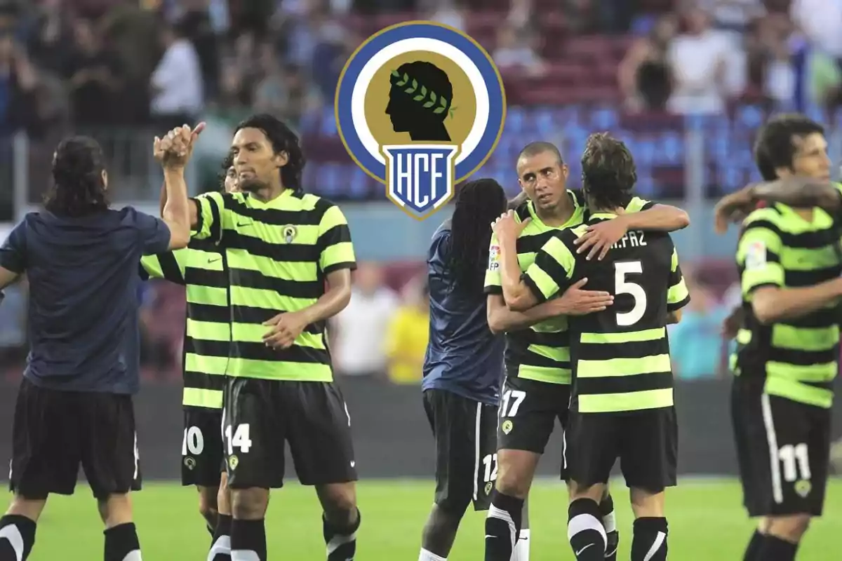 Jugadores de fútbol con uniformes a rayas verdes y negras celebran en el campo con el escudo del Hércules CF en el fondo.