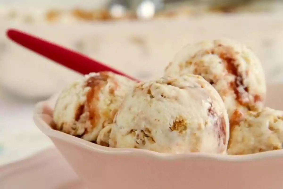 Tres bolas de helado de vainilla con trozos de galleta y caramelo en un tazón rosa con una cuchara roja.