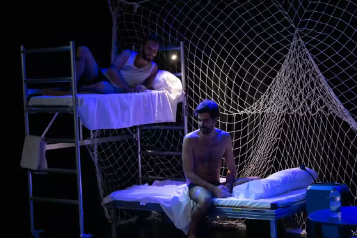 Dos hombres en un escenario teatral con literas y una red de fondo, uno está sentado en la cama inferior y el otro recostado en la cama superior, iluminados con luz azul.