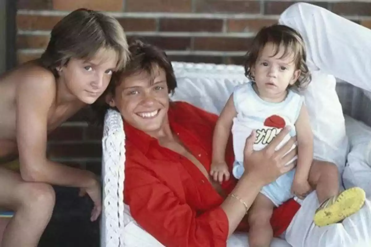 Tres niños posan juntos, uno de ellos con una camisa roja y sosteniendo a un bebé.