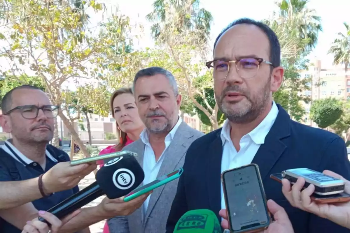 Un grupo de personas, algunas con micrófonos y teléfonos, entrevistando a un hombre con gafas y barba en un entorno al aire libre con árboles y edificios al fondo.