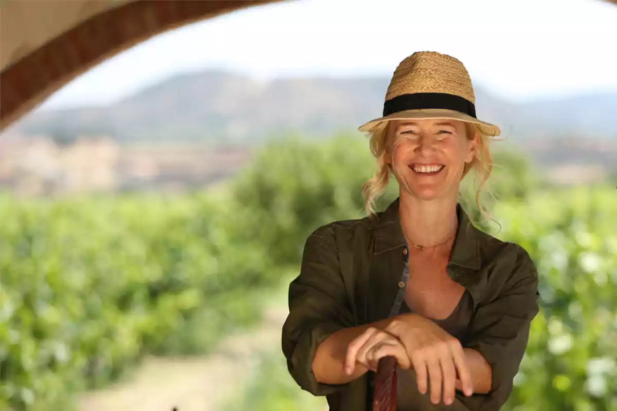 Una mujer sonriente con sombrero de paja y camisa verde, de pie en un entorno natural con vegetación y montañas al fondo.