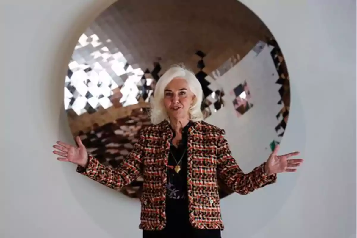 Mujer de cabello blanco con chaqueta colorida posando frente a una obra de arte circular y reflectante.