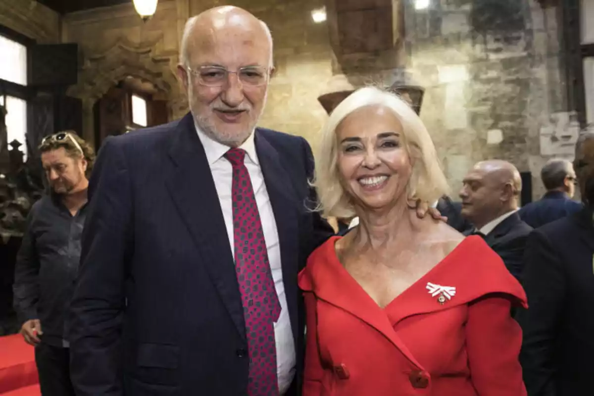 Una pareja sonriente posando en un evento formal, con un hombre de traje y corbata y una mujer con un vestido rojo, rodeados de otras personas en un entorno elegante.