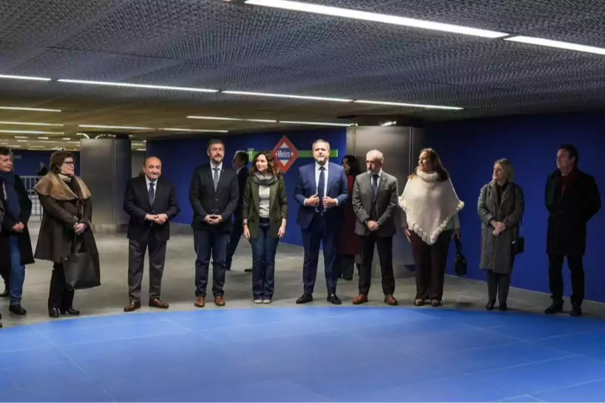 Un grupo de personas de pie en una estación de metro con un fondo azul y un cartel que indica la entrada al metro.