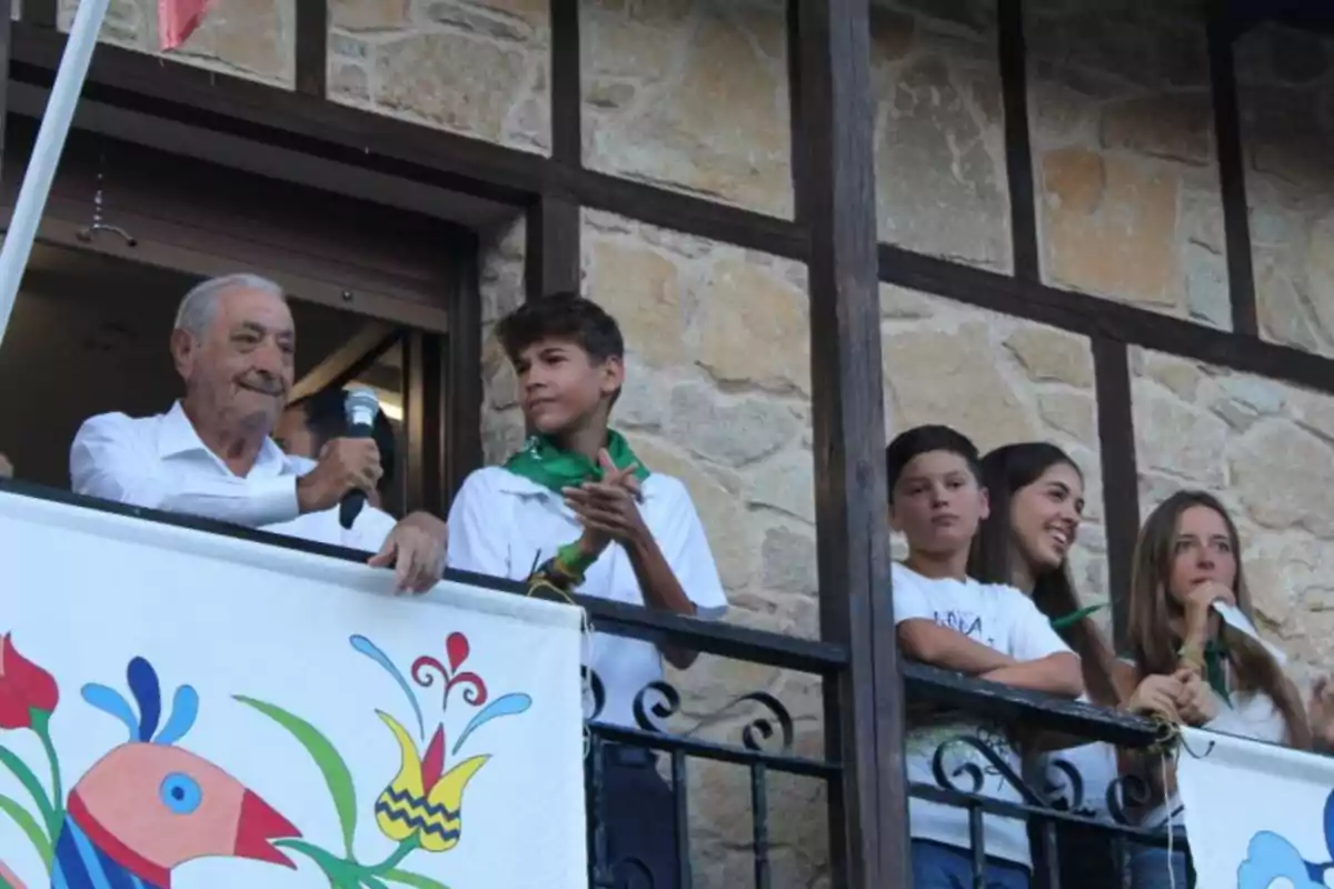 Un hombre mayor con un micrófono y varios jóvenes en un balcón decorado con una pancarta colorida.