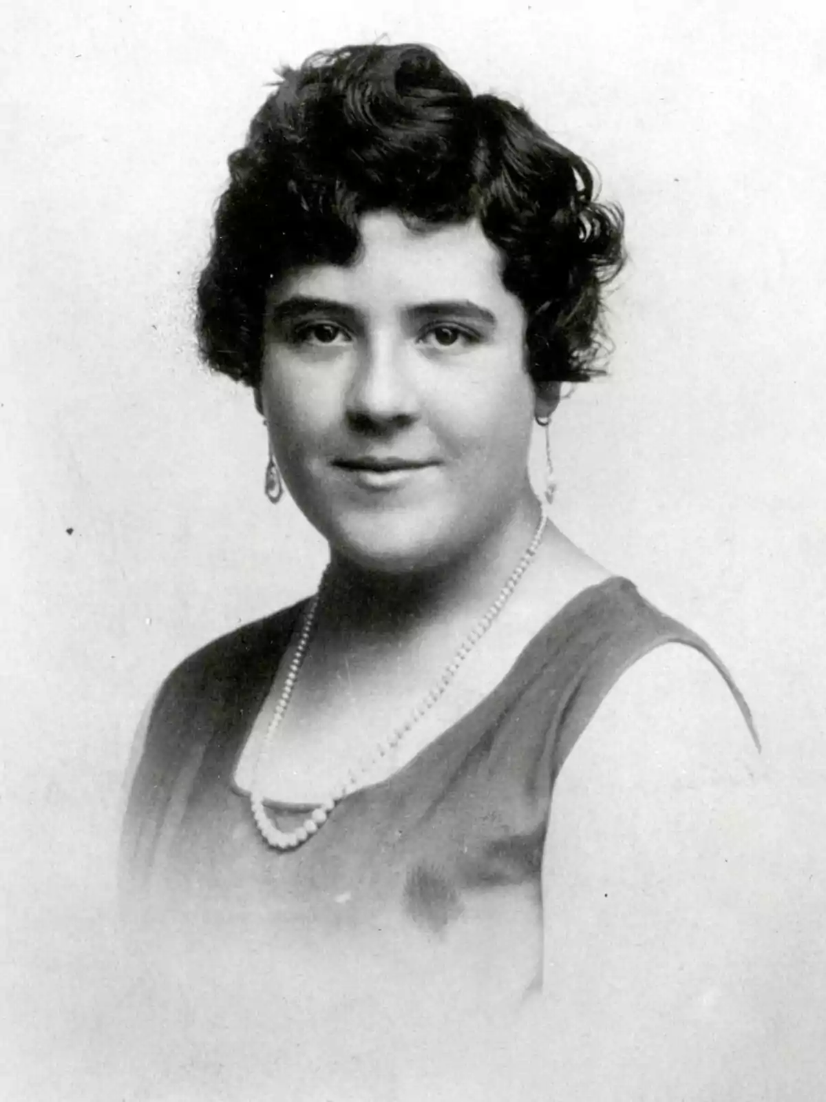 Retrato en blanco y negro de una mujer joven con cabello corto y rizado, usando un vestido oscuro y un collar de perlas.