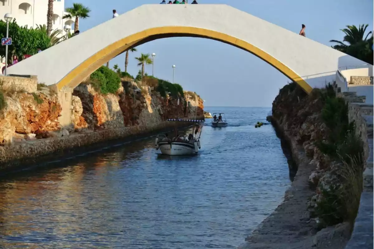 Un puente blanco con borde amarillo cruza un canal de agua donde navegan pequeñas embarcaciones rodeadas de paredes rocosas y vegetación Un puente blanco con borde amarillo cruza un canal de agua donde navegan pequeñas embarcaciones rodeadas de paredes rocosas y vegetación