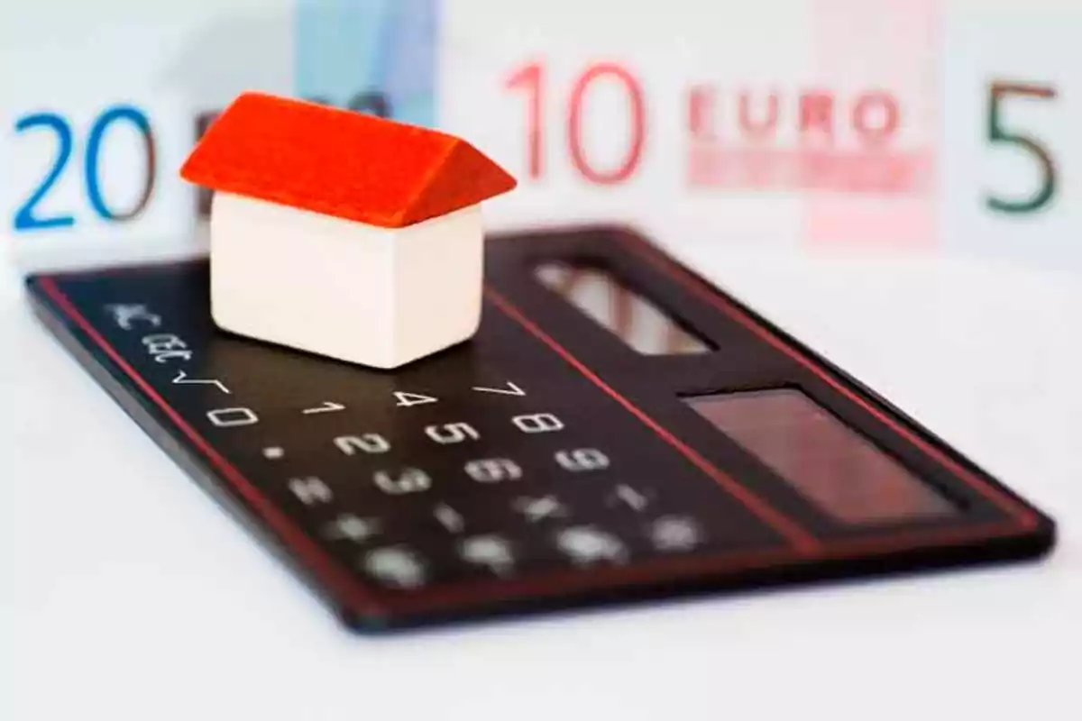 Casa en miniatura sobre una calculadora con billetes de euro al fondo.