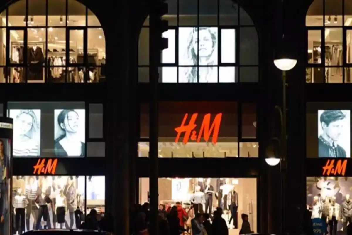 Fachada de una tienda H&M iluminada por la noche con maniquíes y carteles publicitarios en las ventanas.