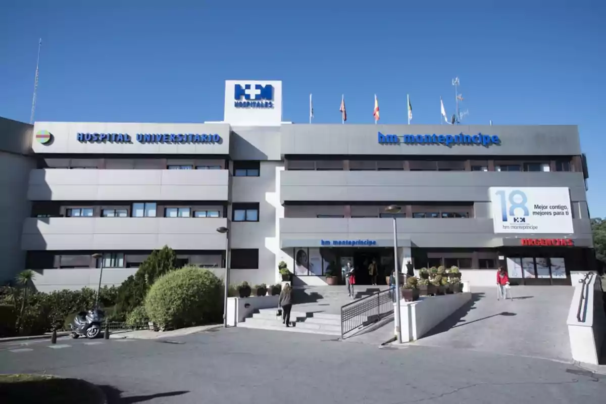 Fachada de un hospital universitario con varias personas entrando y saliendo, rodeado de vegetación y con un cielo despejado.