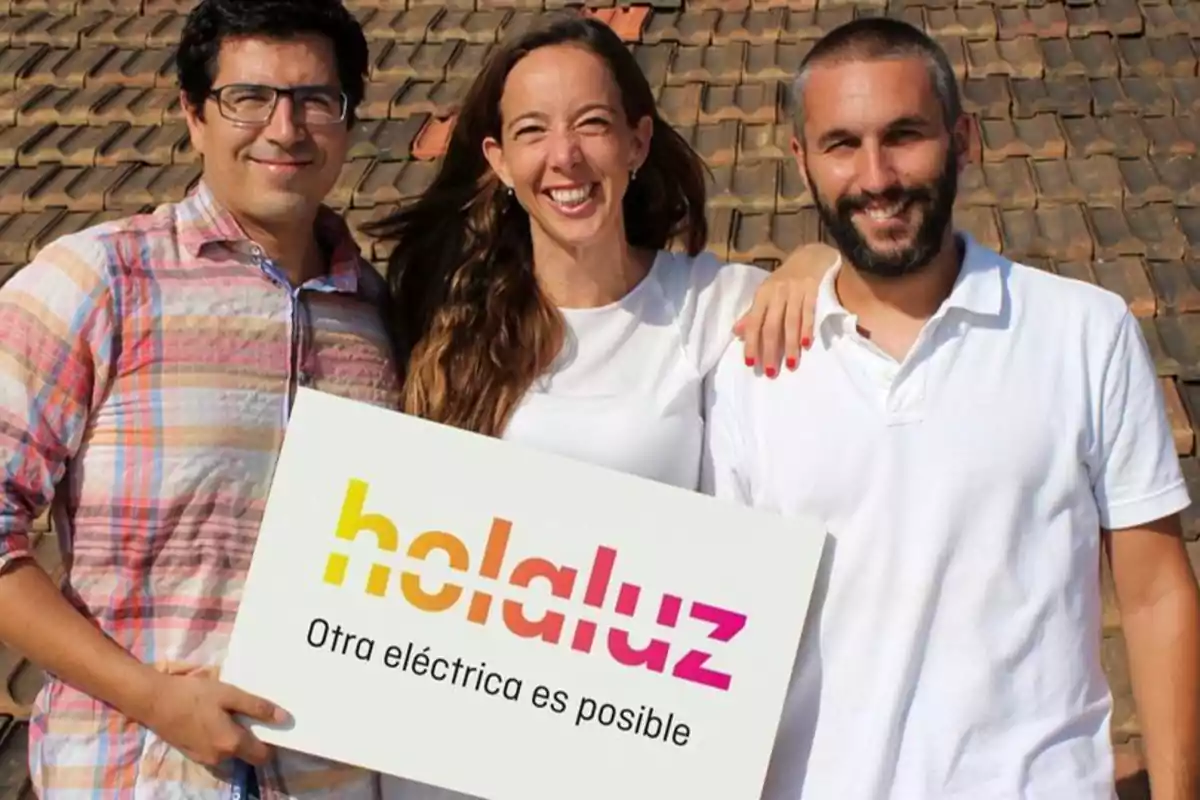 Tres personas sonrientes sostienen un cartel que dice "holaluz Otra eléctrica es posible" frente a un fondo de tejas.