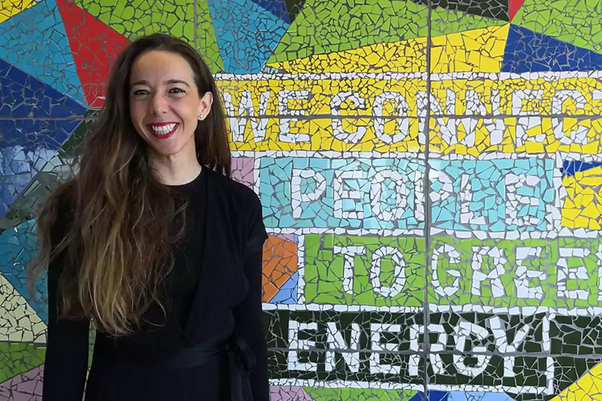 Una mujer sonriente con cabello largo y suelto está de pie frente a un colorido mural de mosaico que tiene un mensaje en inglés sobre conectar personas con energía verde.