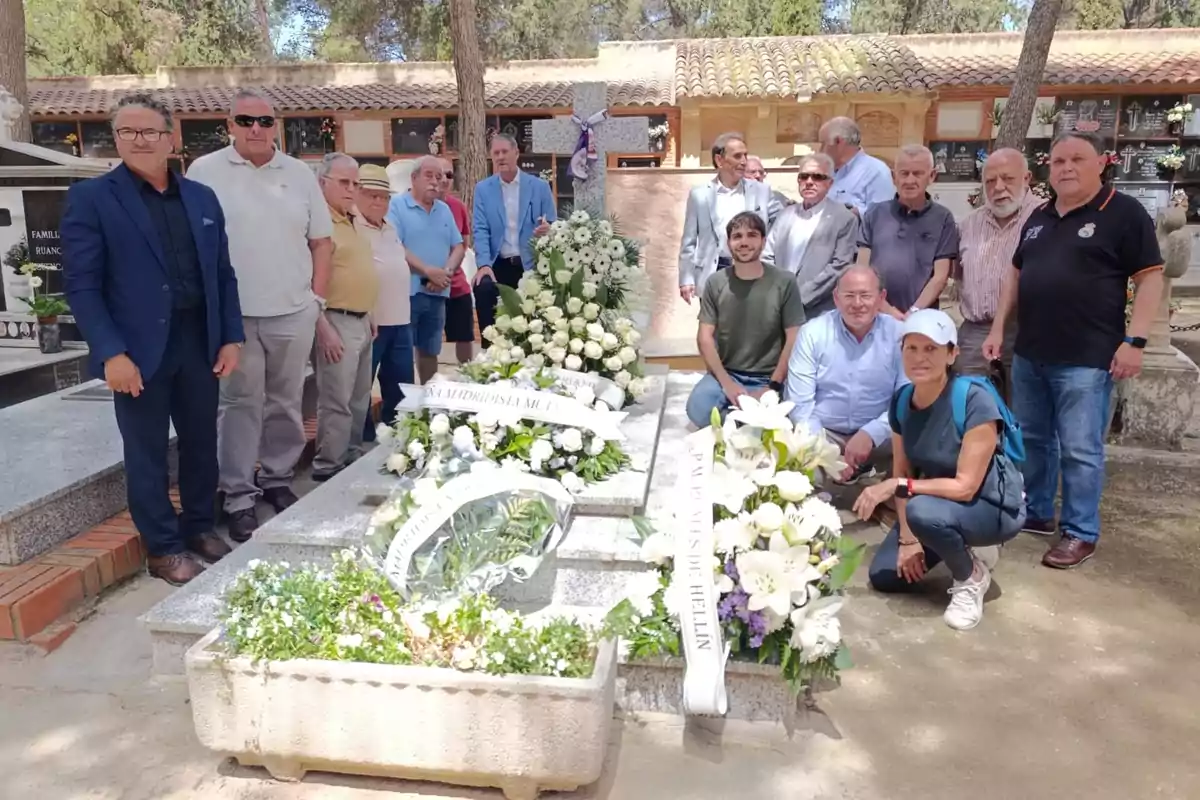 Un grupo de personas posa junto a una tumba adornada con arreglos florales en un cementerio durante el día.