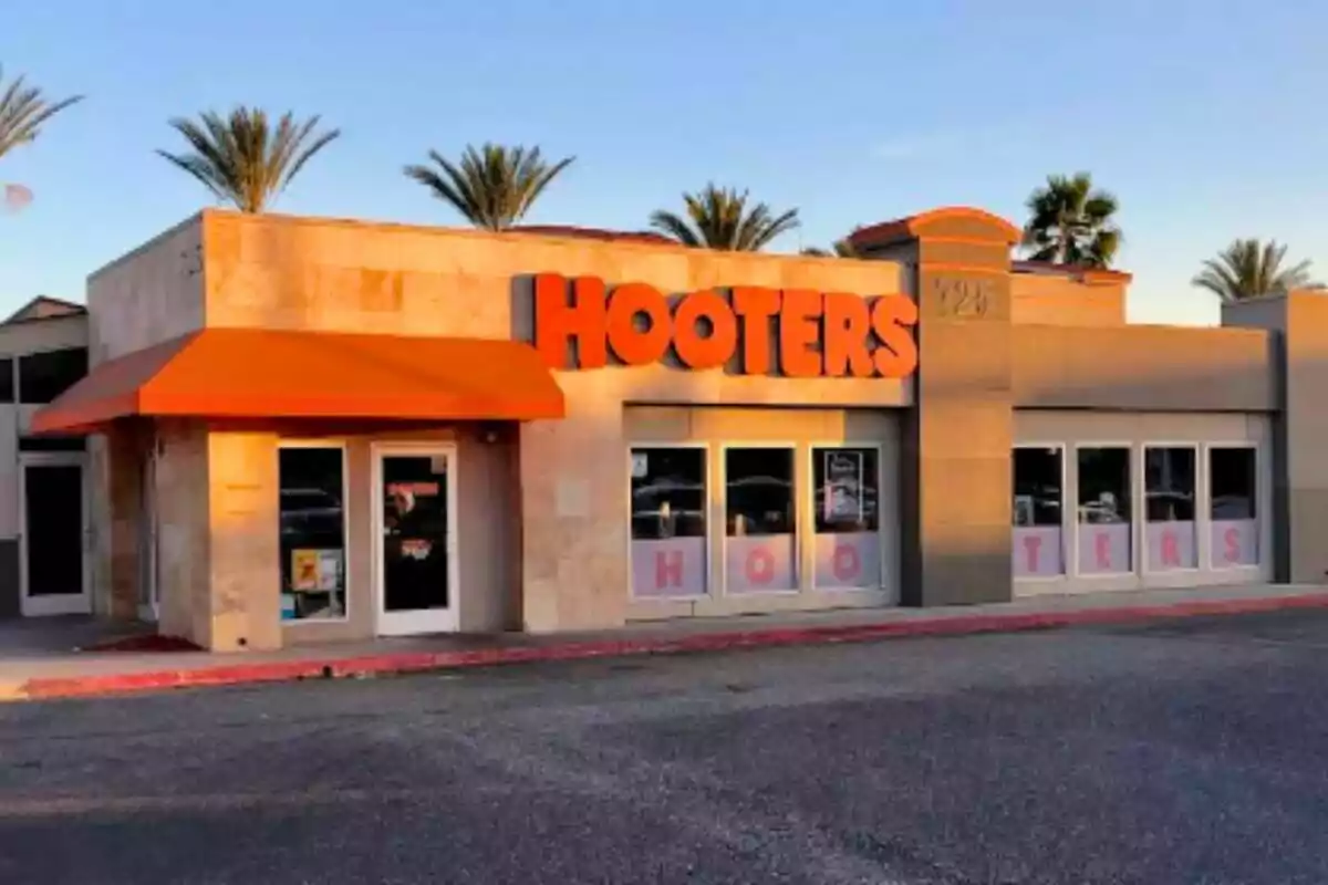 Fachada de un restaurante Hooters con toldo naranja y palmeras al fondo Fachada de un restaurante Hooters con toldo naranja y palmeras al fondo