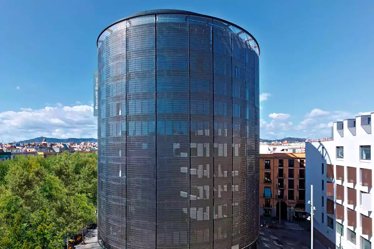 Edificio cilíndrico de cristal oscuro en un entorno urbano con cielo despejado.